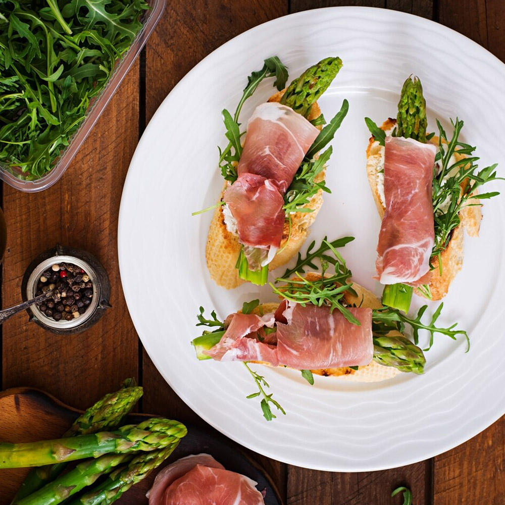 Prosciutto wrapped around asparagus on top of bread on a plate next to a jar of peppercorns and other vegetables