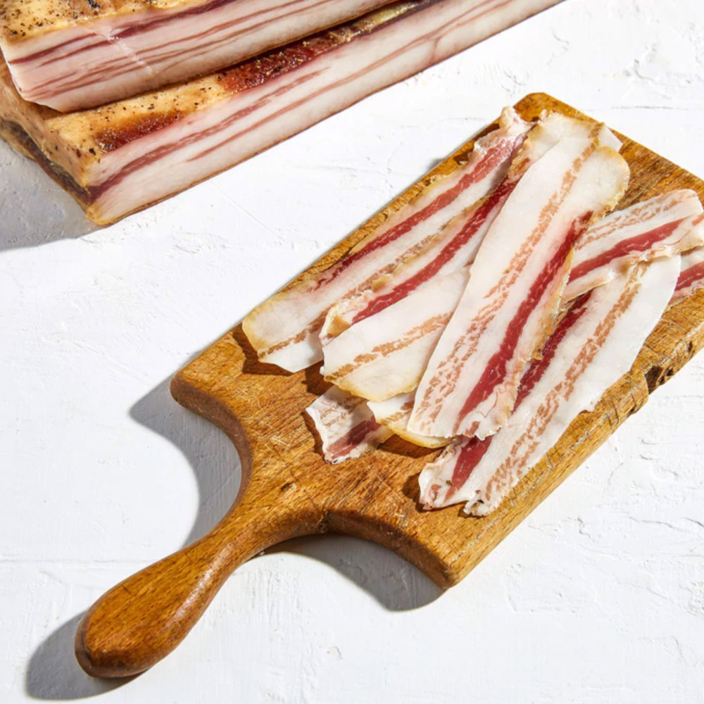 Slices of pancetta on a wood board next to a slab of pancetta