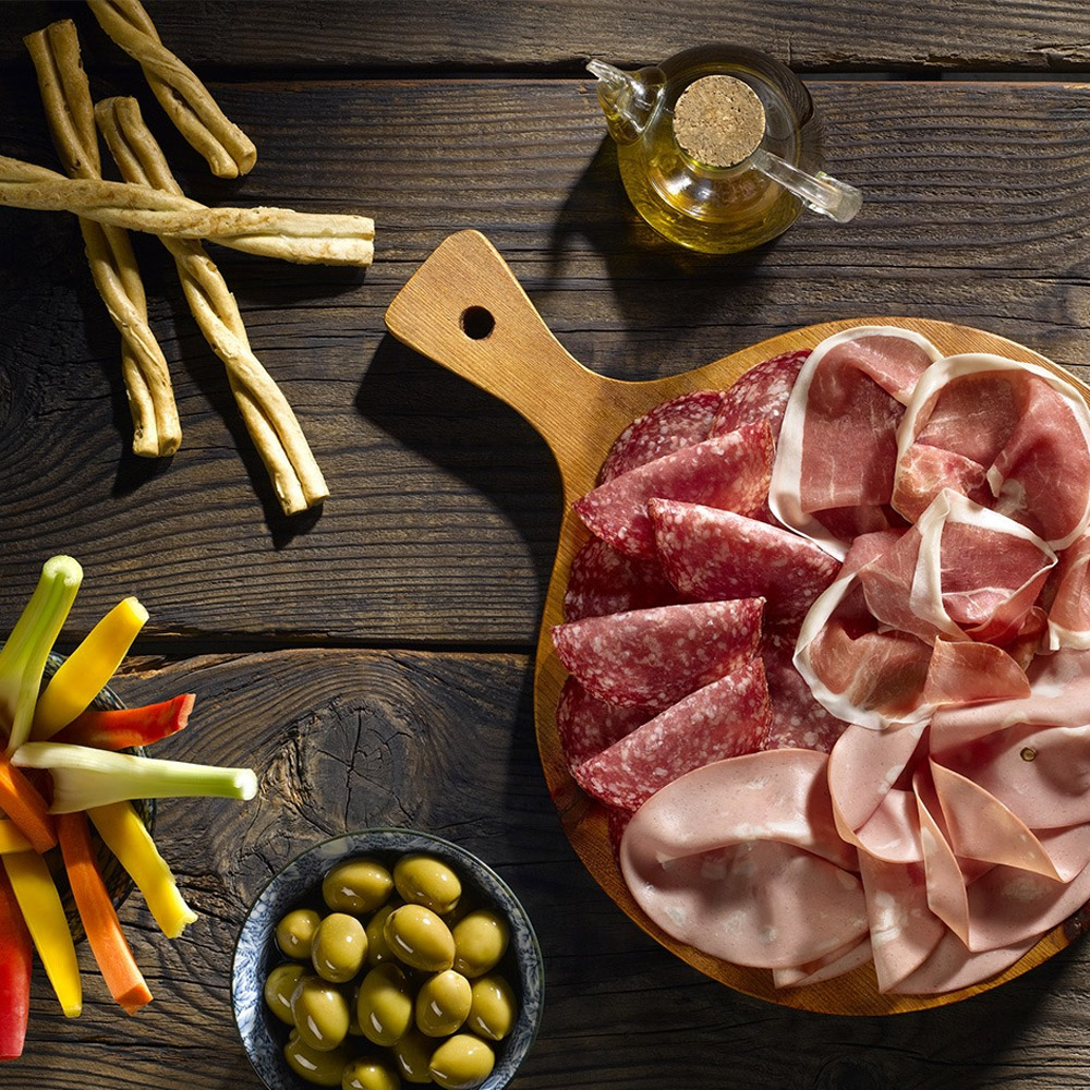 A wood board with salami and prosciutto next to a bowl of olives and some dried breadsticks
