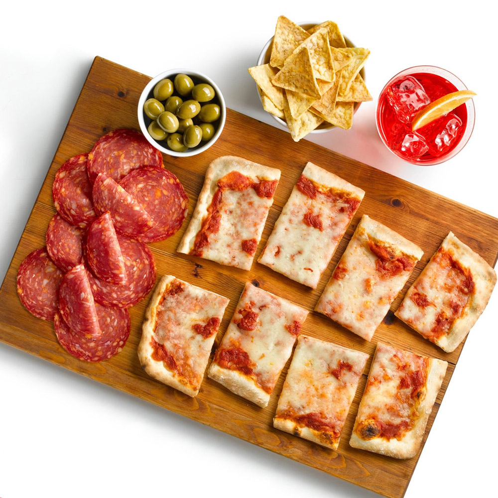 Slices of salami on a wood board with pieces of pizza and a bowl of olives next to a bowl of chips and a cocktail
