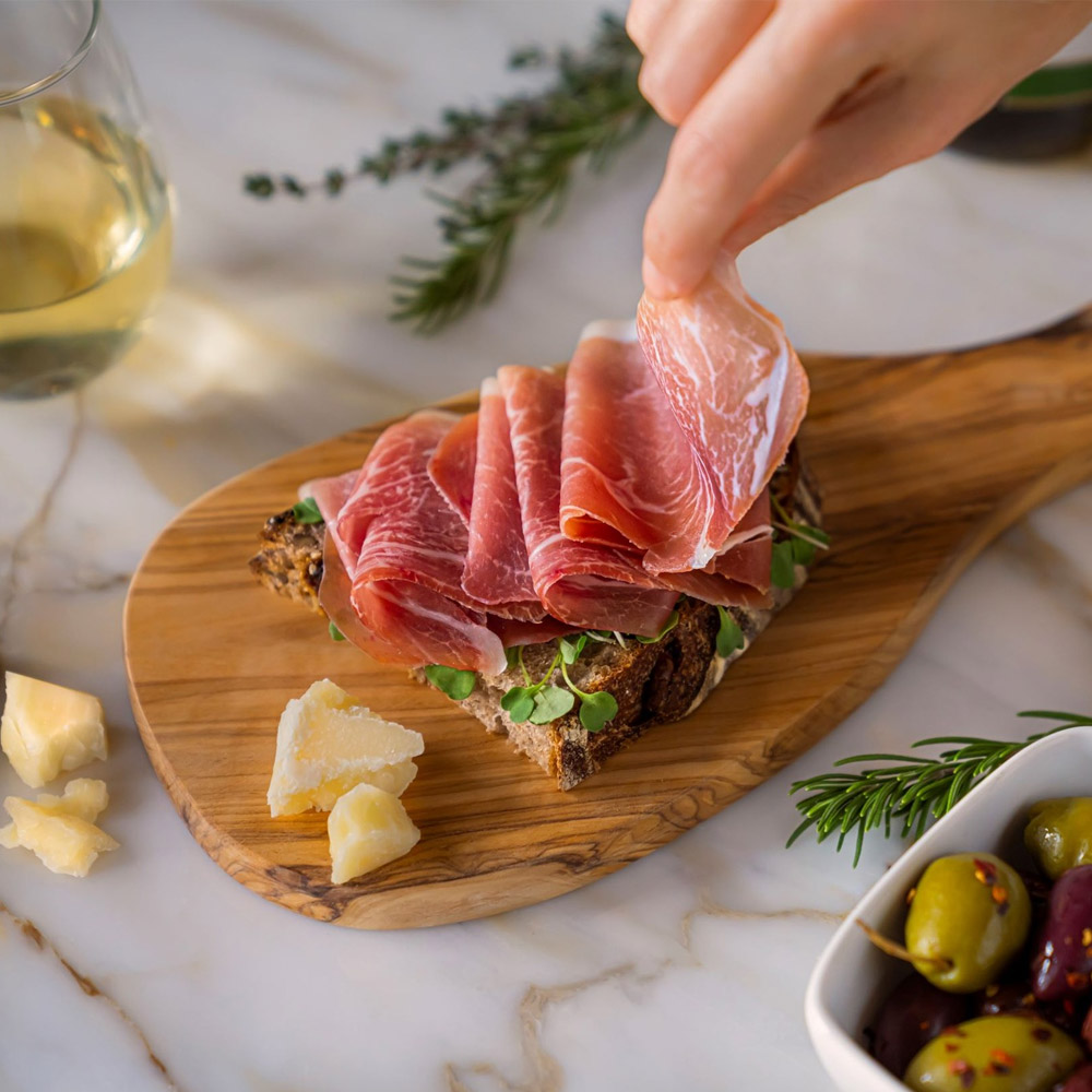A slice of prosciutto on a wood board next to a glass of wine and a bowl of olives
