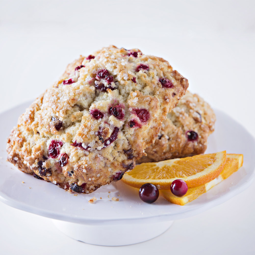 Sweet Street artisan cranberry orange scones on a white cake stand with fresh oranges and cranberries