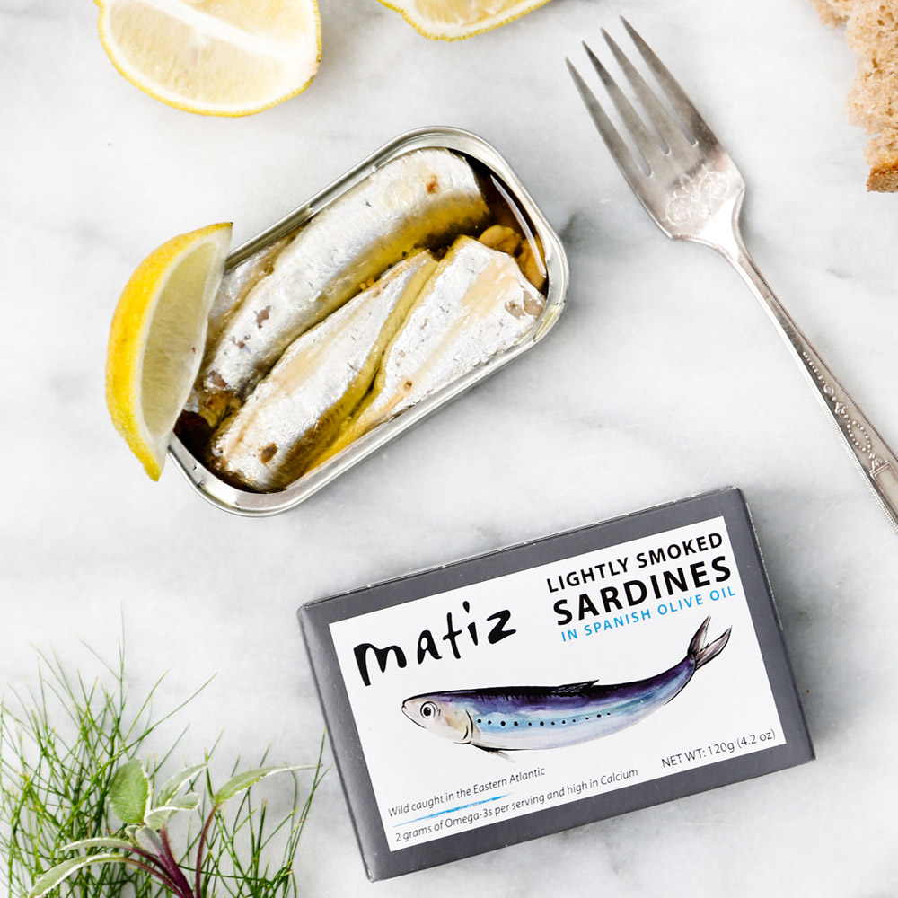 An open tin of Matiz Lightly Smoked Sardines In Olive Oil pictured with the box packaging and a fork and lemon wedge