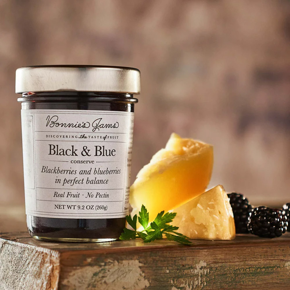 A jar of Bonnie's Jams Black & Blue on a wood board next to cheese and blackberries