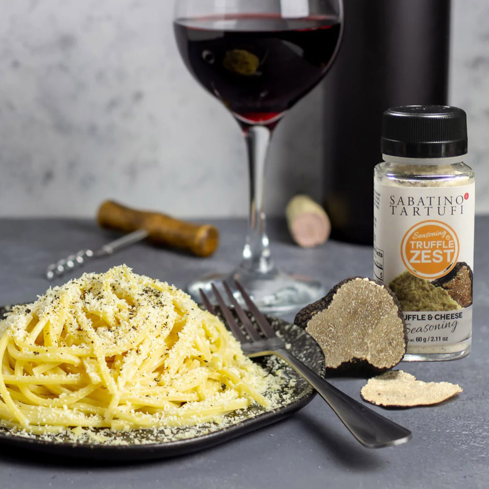 A jar of Sabatino Tartufi Truffle & Cheese Seasoning next to a plate of pasta and a truffle with a glass of wine