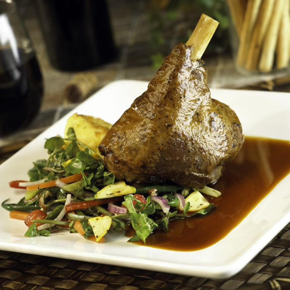 A lamb shank on a plate surrounded with salad and lamb stock