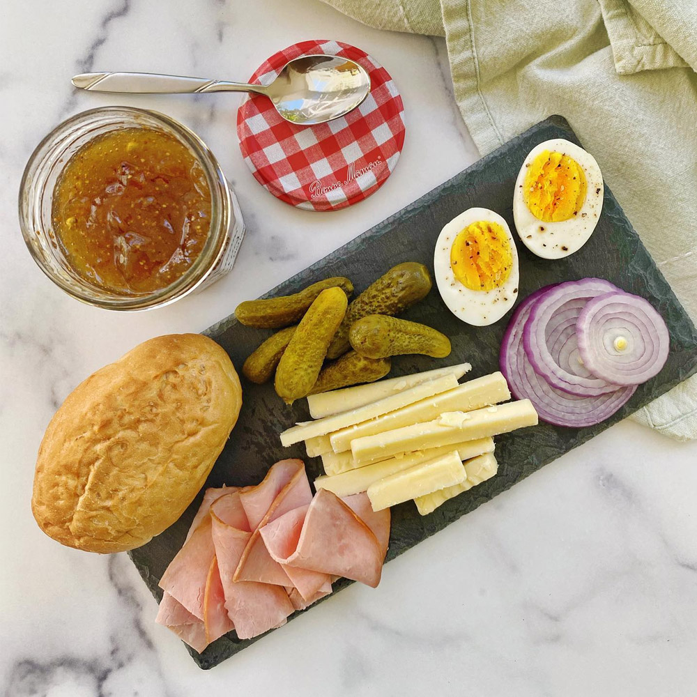 Cheese on a wood board with onion, pickles, ham, hard boiled egg and a dinner roll next to an open jar of jam