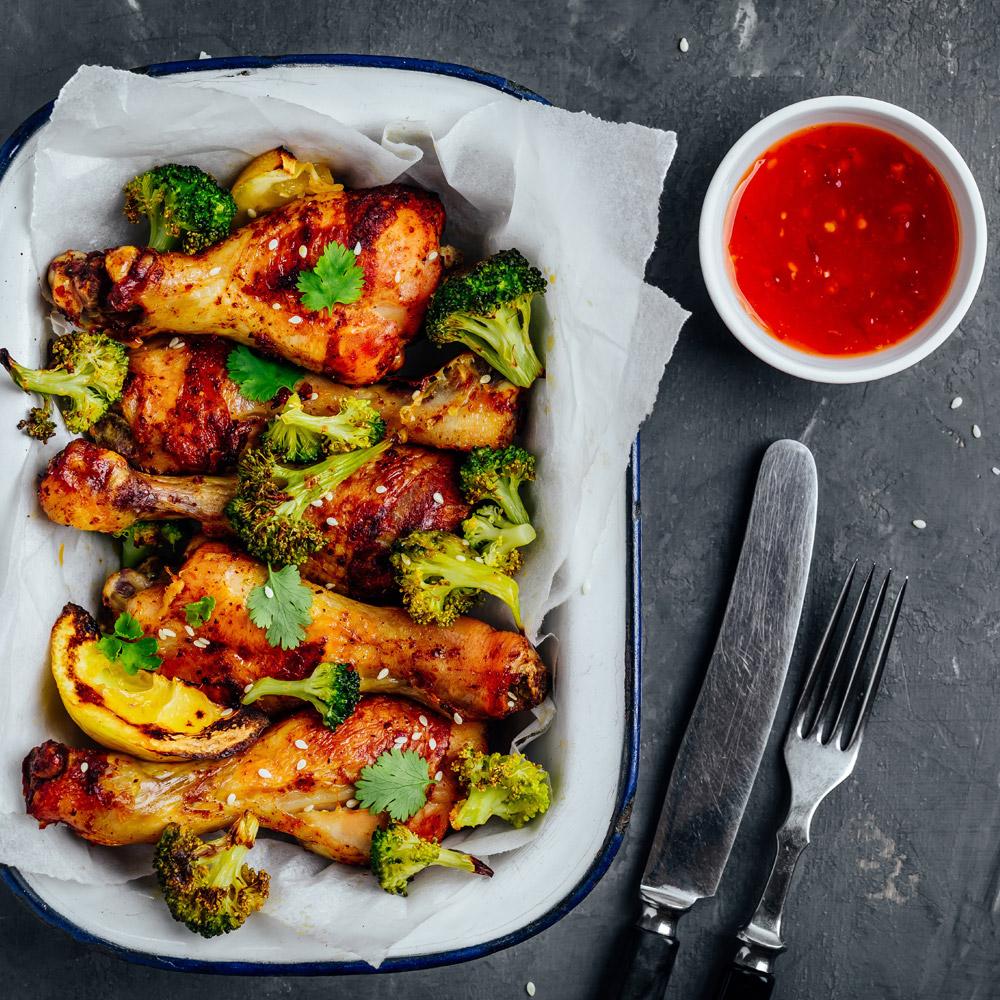Baked chicken drumsticks with broccoli cilantro sesame and sweet chili sauce on a gray stone background