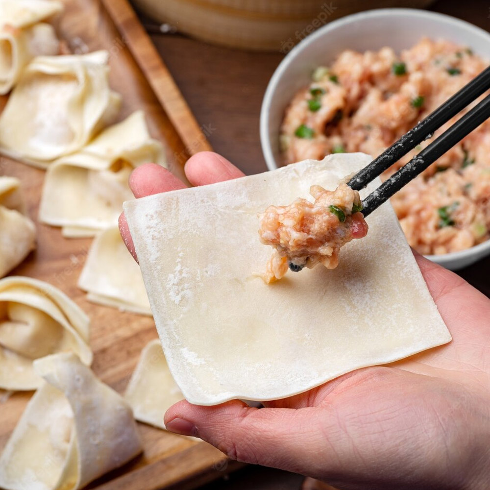 A hand holding a wonton wrapper next to a bowl of pork filling and a board of formed wontons