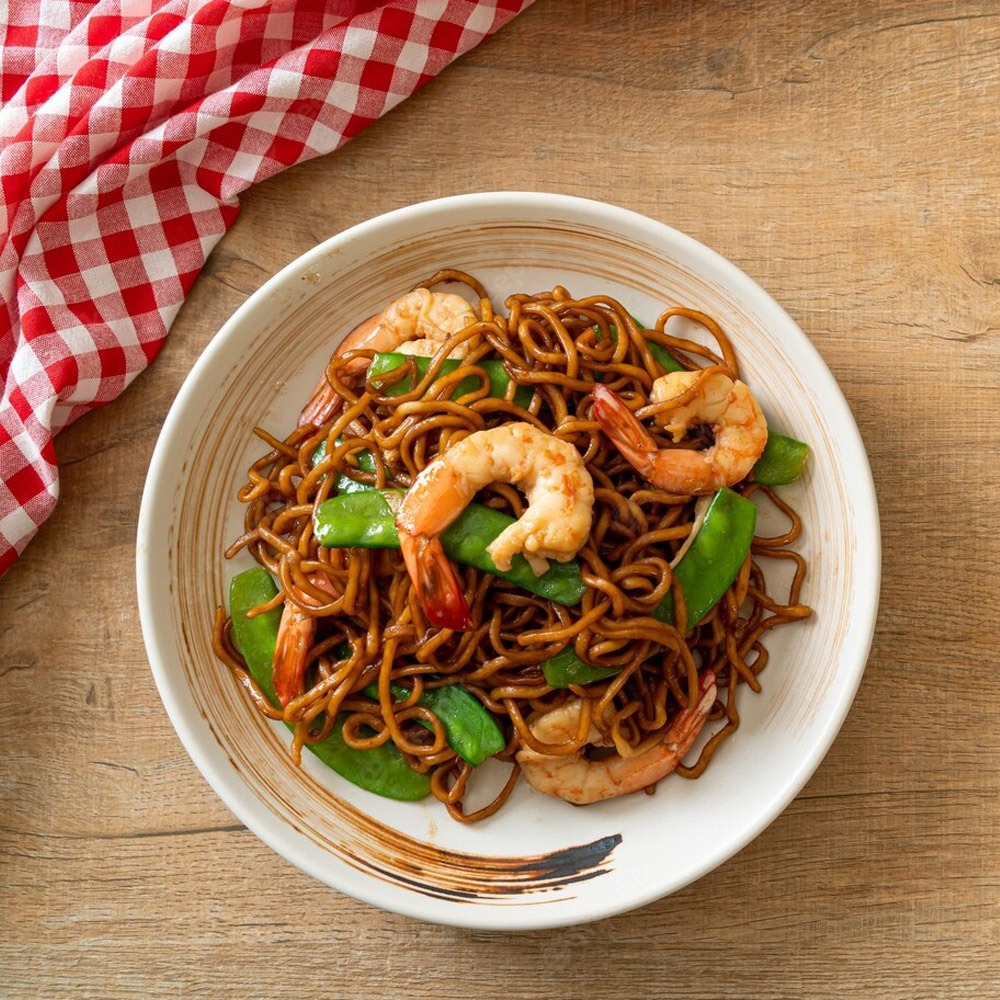 Bowl of cooked soba noodles with shrimp and snow peas on a table next to a scrunched table cloth