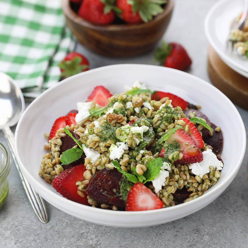 A bowl of farro salad next to a spoon and a bowl of strawberries