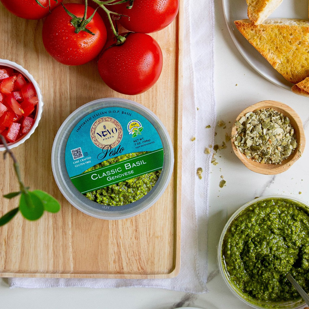 A container of Nuovo Classic Basil Pesto on a wood board with tomatoes next to a bowl of pesto