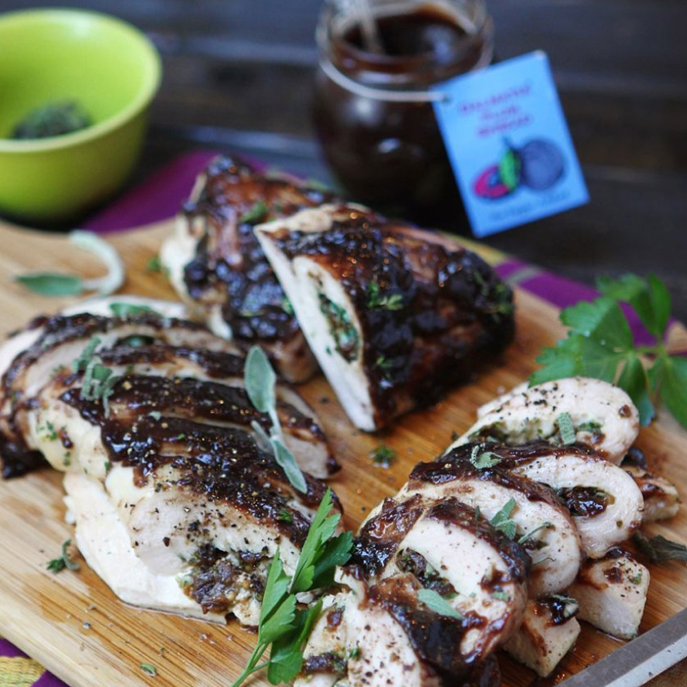 Slices of grilled chicken topped with Dalmatia Plum Spread on a wood board in front of an open jar of Dalmatia Plum Spread