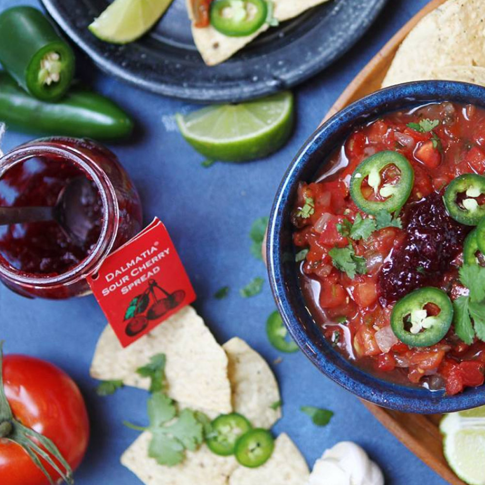 An open jar of Dalmatia Sour Cherry Spread next to a bowl of salsa topped with jalapenos and sour cherry spread