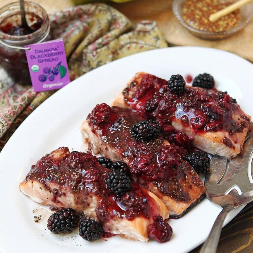 A plate of grilled salmon topped with blackberry spread with an open jar of Dalmatia Blackberry Spread in the background