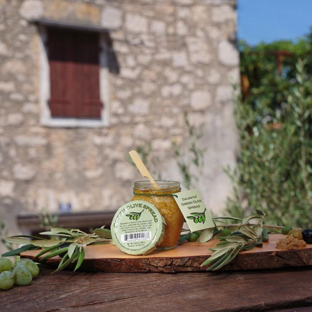 An open jar of Dlamatia Green Olive Spread on a wood plank in front of an old building outside