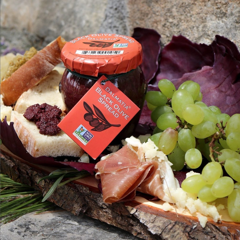 A jar of Dalmatia Black Olive spread on a board with fruit and prosciutto next to a piece of bread with black olive spread on top