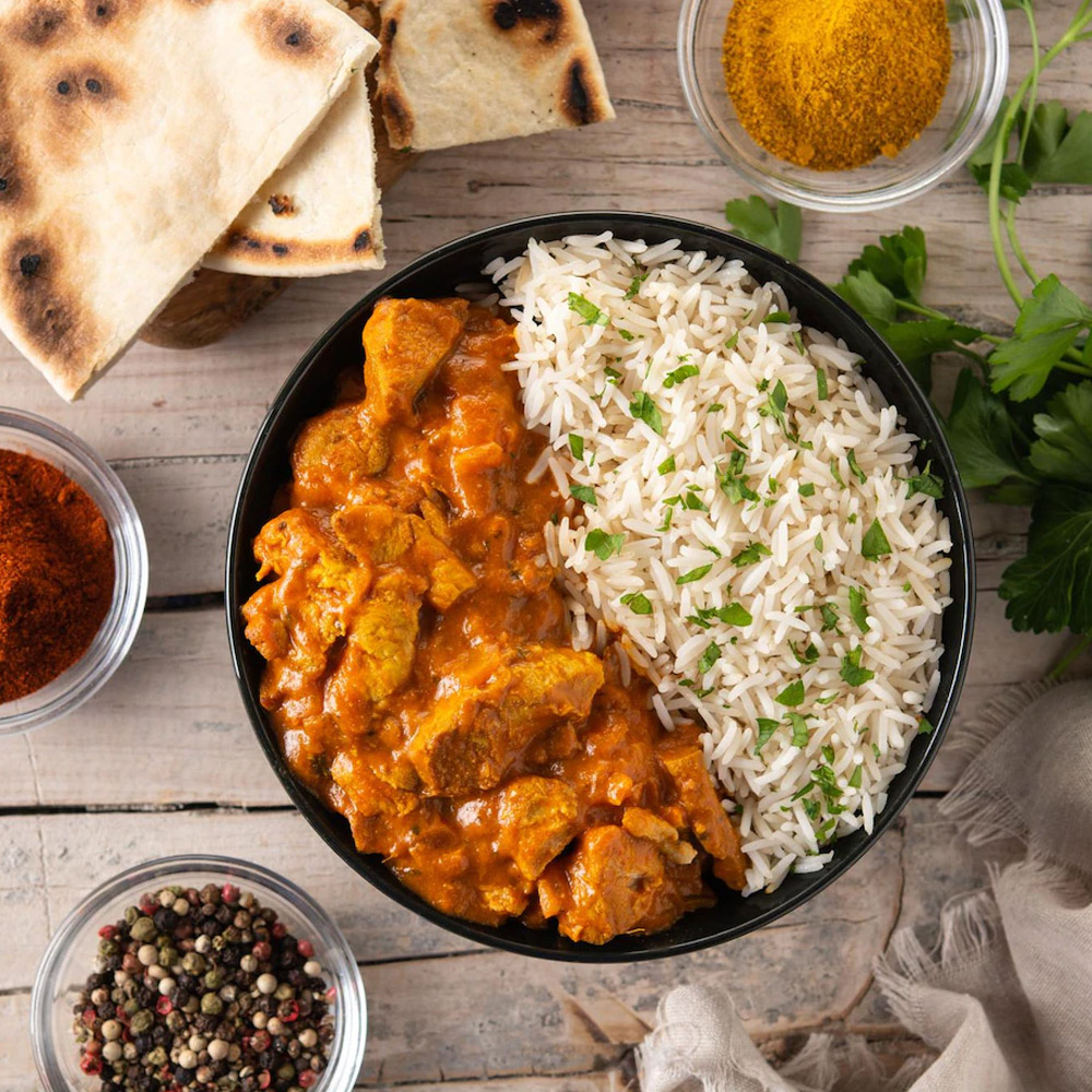 Indian butter chicken and rice in black bowl on wooden table next to spices and pita bread