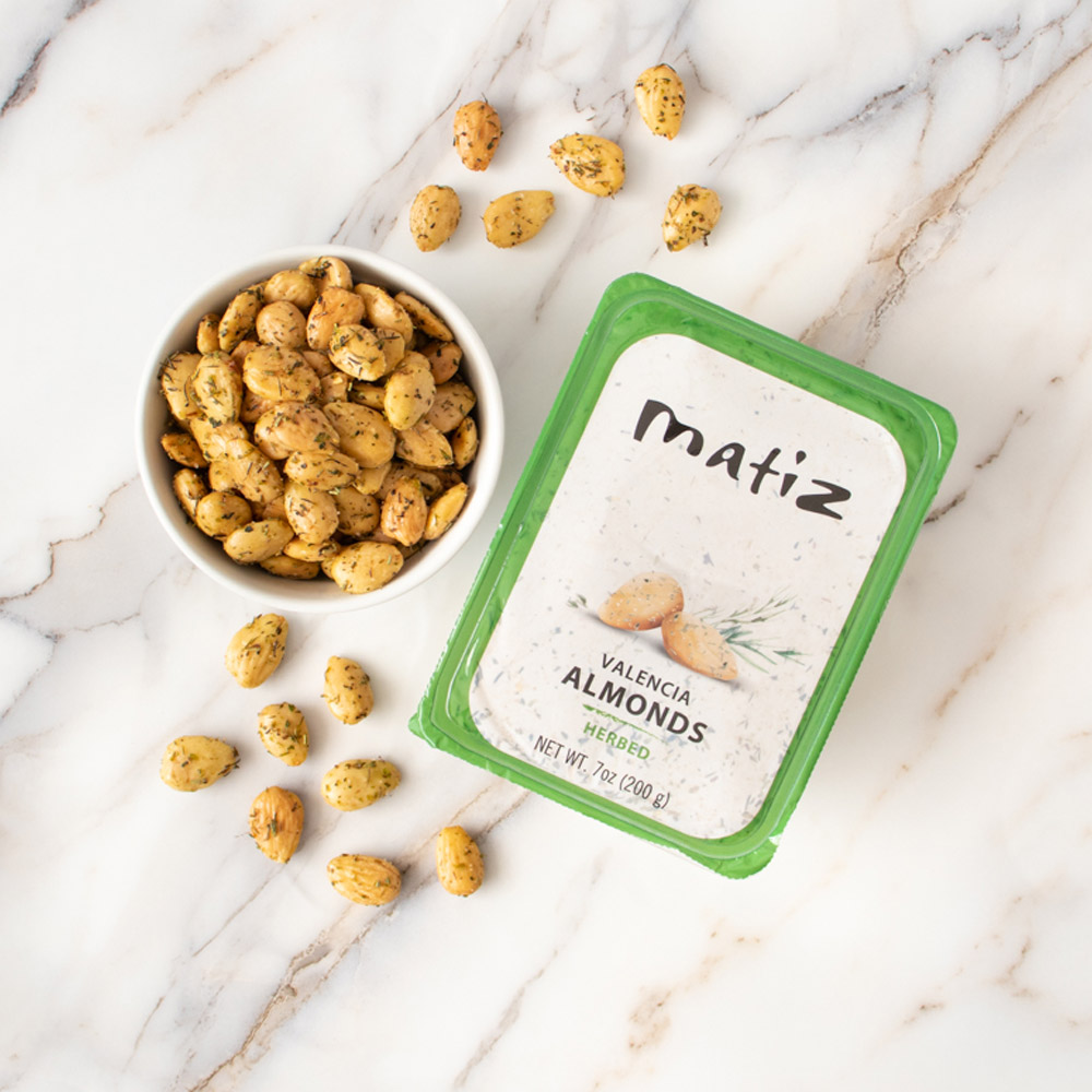 A container of Matiz Valencia Almonds with Herbs next to a bowl of Matiz Valencia Almonds with Herbs