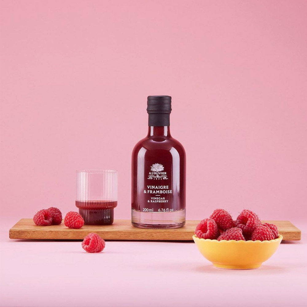 A bottle of A L'Olivier Raspberry Fruit Vinegar next to a glass of vinegar and a bowl of raspberries
