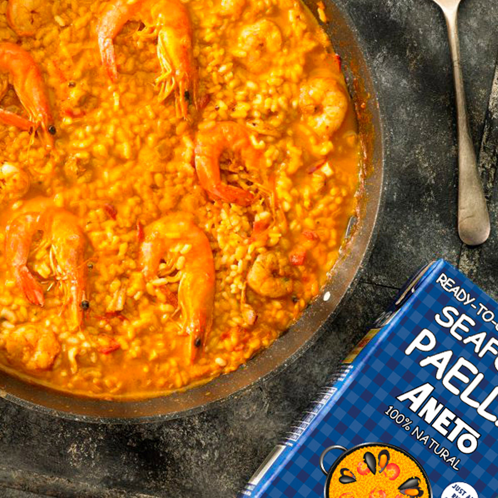 A box of Aneto Seafood paella cooking base laying next to a pan filled with paella