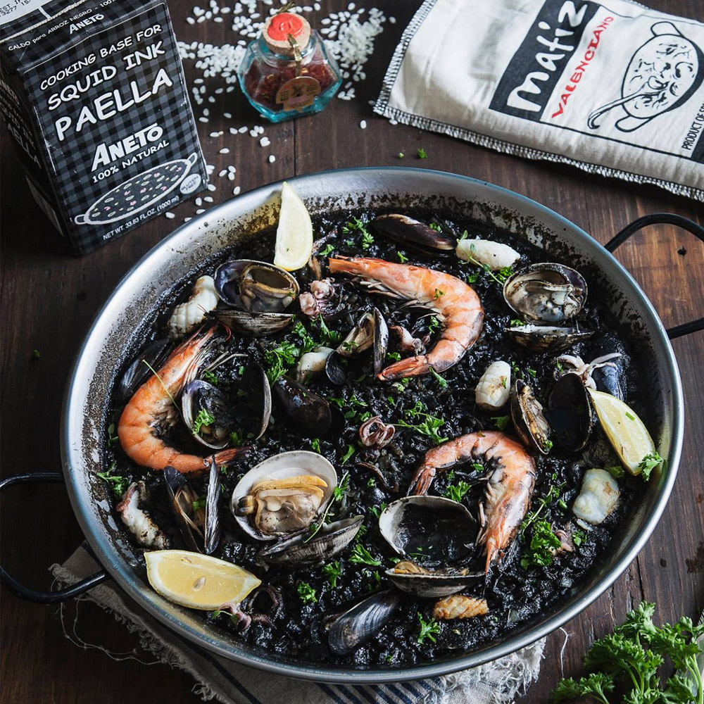 A box of Aneto squid ink paella cooking base next to a pan of squid ink paella and a spilled bag of rice