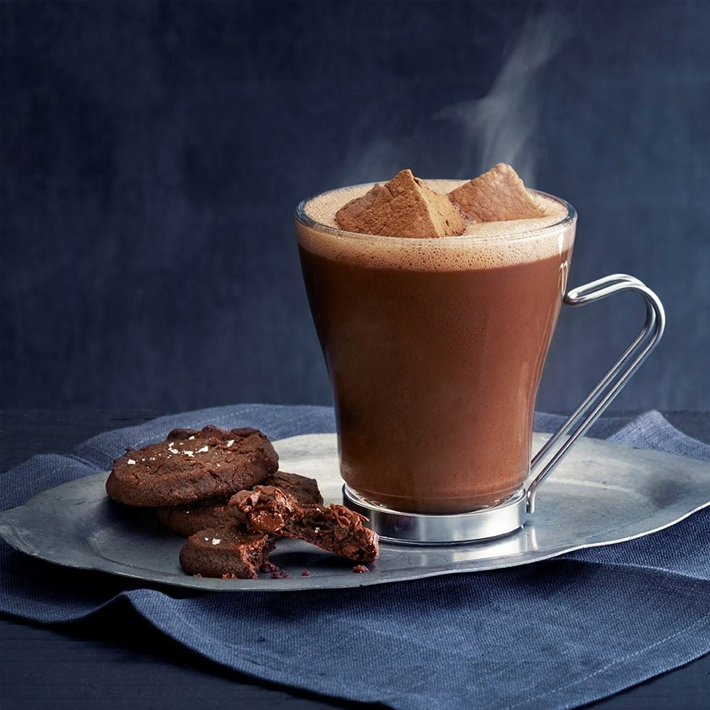A glass mug of hot cocoa next to two chocolate cookies