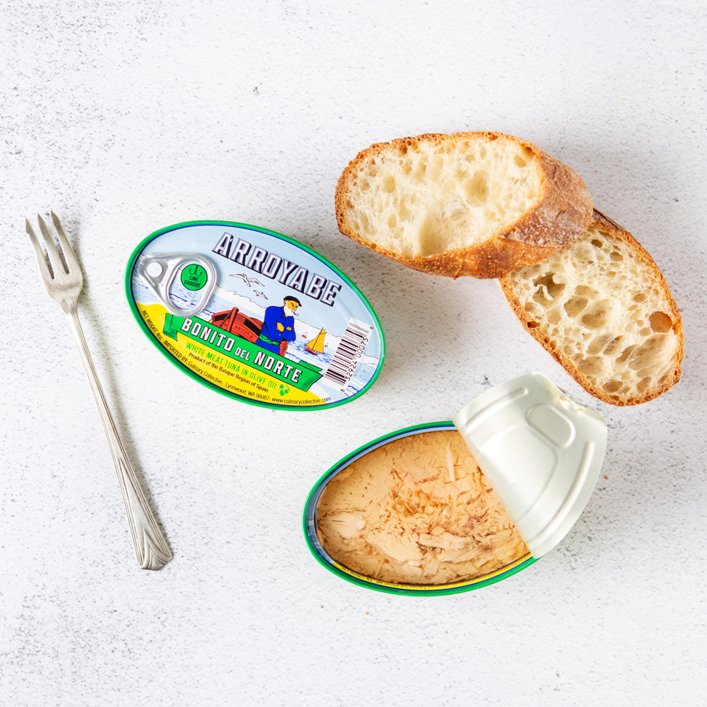 An open tin of Arroyabe Bonito Del Norte in Olive Oil next to a closed tin with a fork and twi pieces of crusty bread