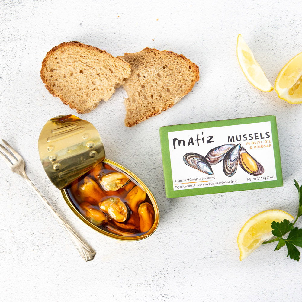 An open tin of Matiz Mussels in Olive Oil & Vinegar pictured with the box packaging and some bread and a fork
