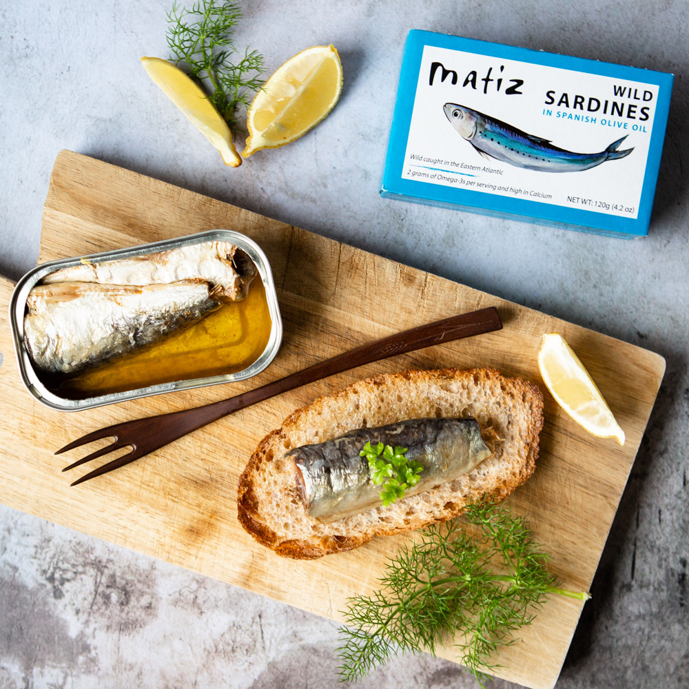 A box of Matiz wild sardines in Spanish olive oil next to a wood board with an open tin of sardines and a fork with a sardine crostini and lemon wedges
