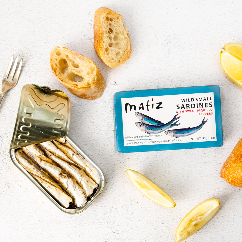 An open tin of Matiz wild small sardines with sweet piquillo peppers pictured with the box packaging next to two pieces of bread and lemon wedges with a fork