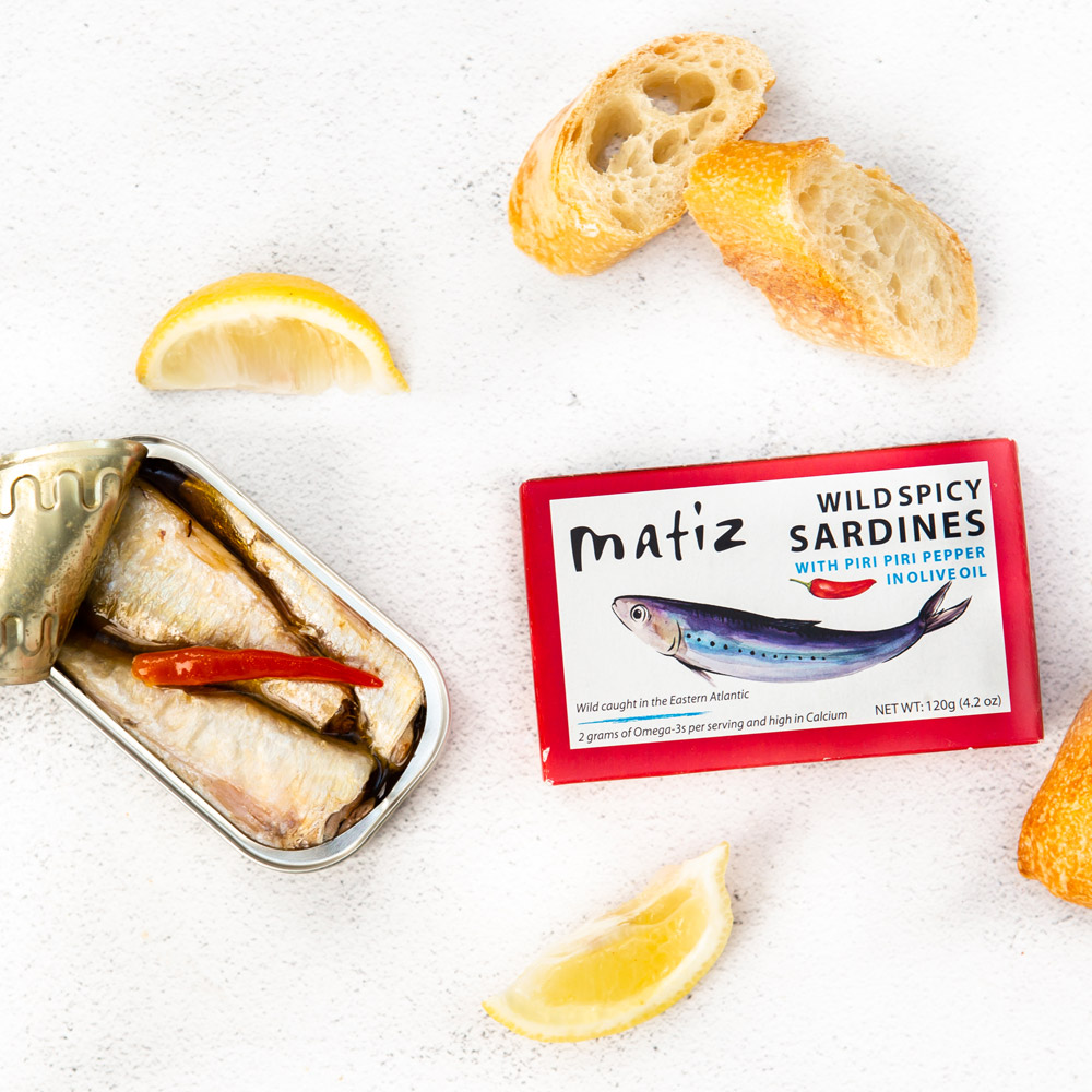 An open tin of Matiz wild spicy sardines with piri piri pepper in olive oil pictured with the box packaging and slices of bread with lemon wedges