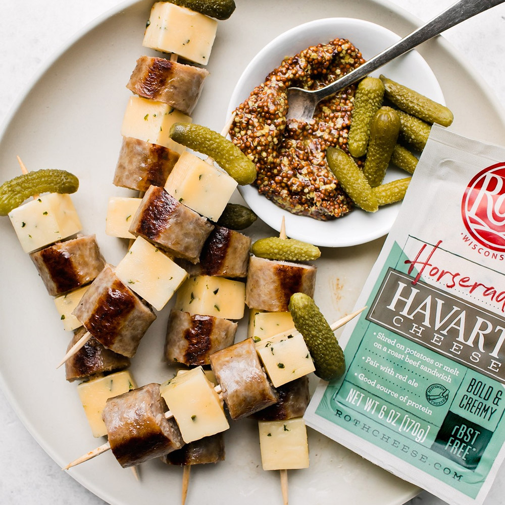 A plate with a bowl of whole grain mustard and havarto, pickle and sausage skewers