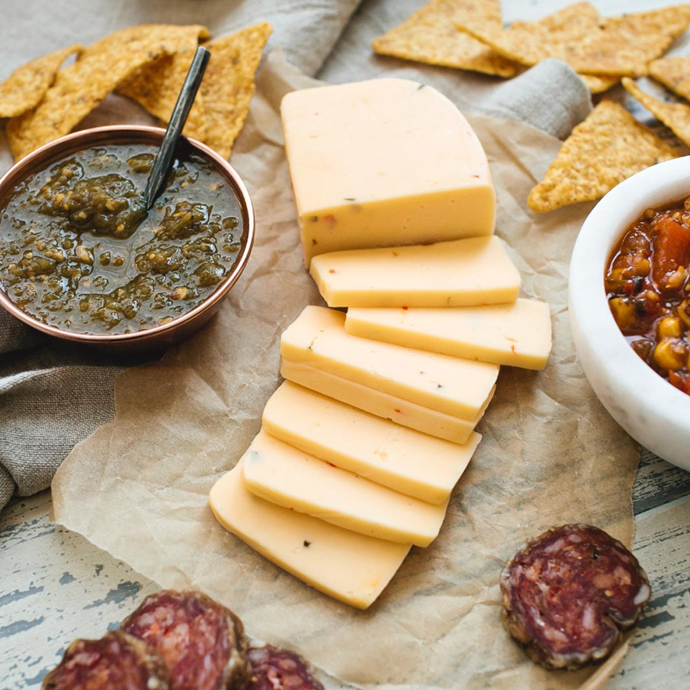 A block of jalapeno havarti next to some salami, tortilla chips and a bowl of salsa
