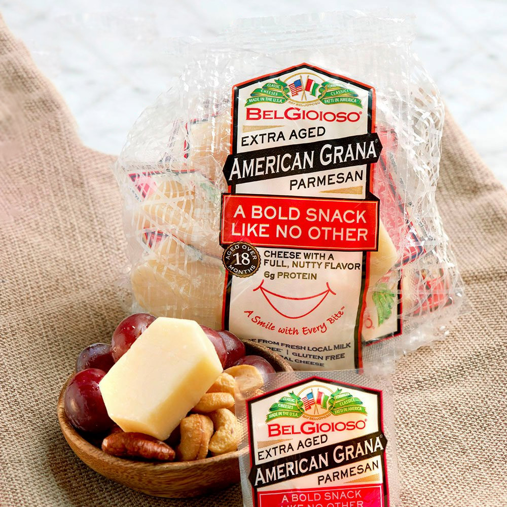 A bag of BelGioioso american grana extra aged parmesan snack packs ehind a small bowl of mixed nuts and fruit