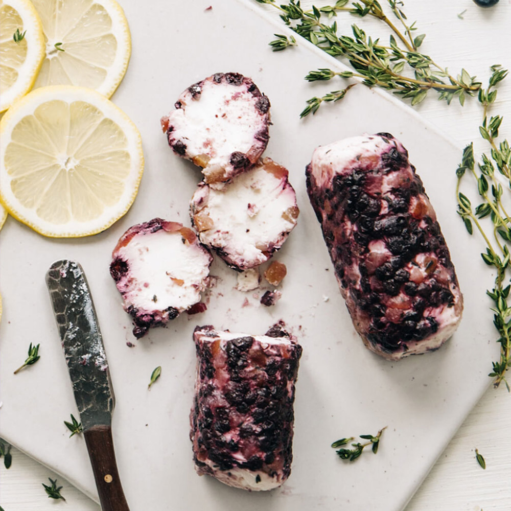 Two blueberry covered goat cheese logs on a cheese board next to slices of lemon