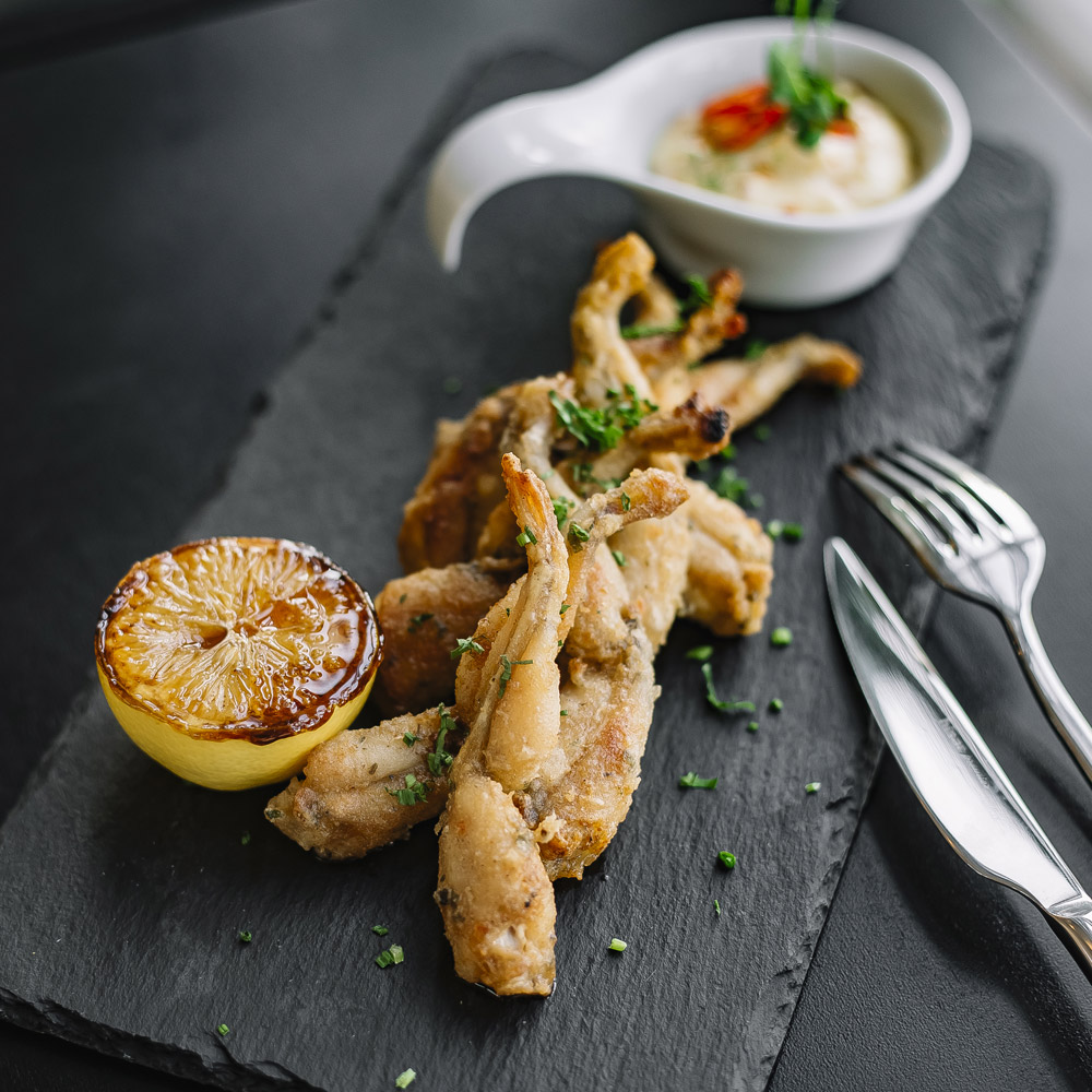Deep fried frog legs on a slate board with a grilled lemon and a bowl of dipping sauce